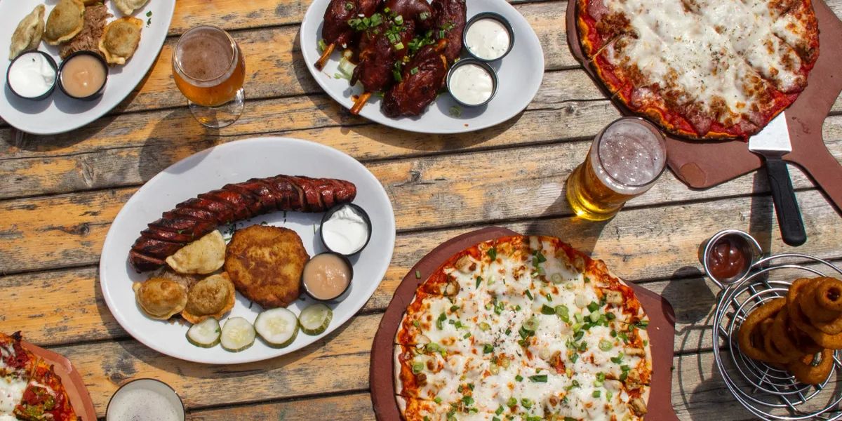 A spread of pizza, pierogi, kielbasa, wings, and beer on a wooden table at Flo & Santos in Chicago