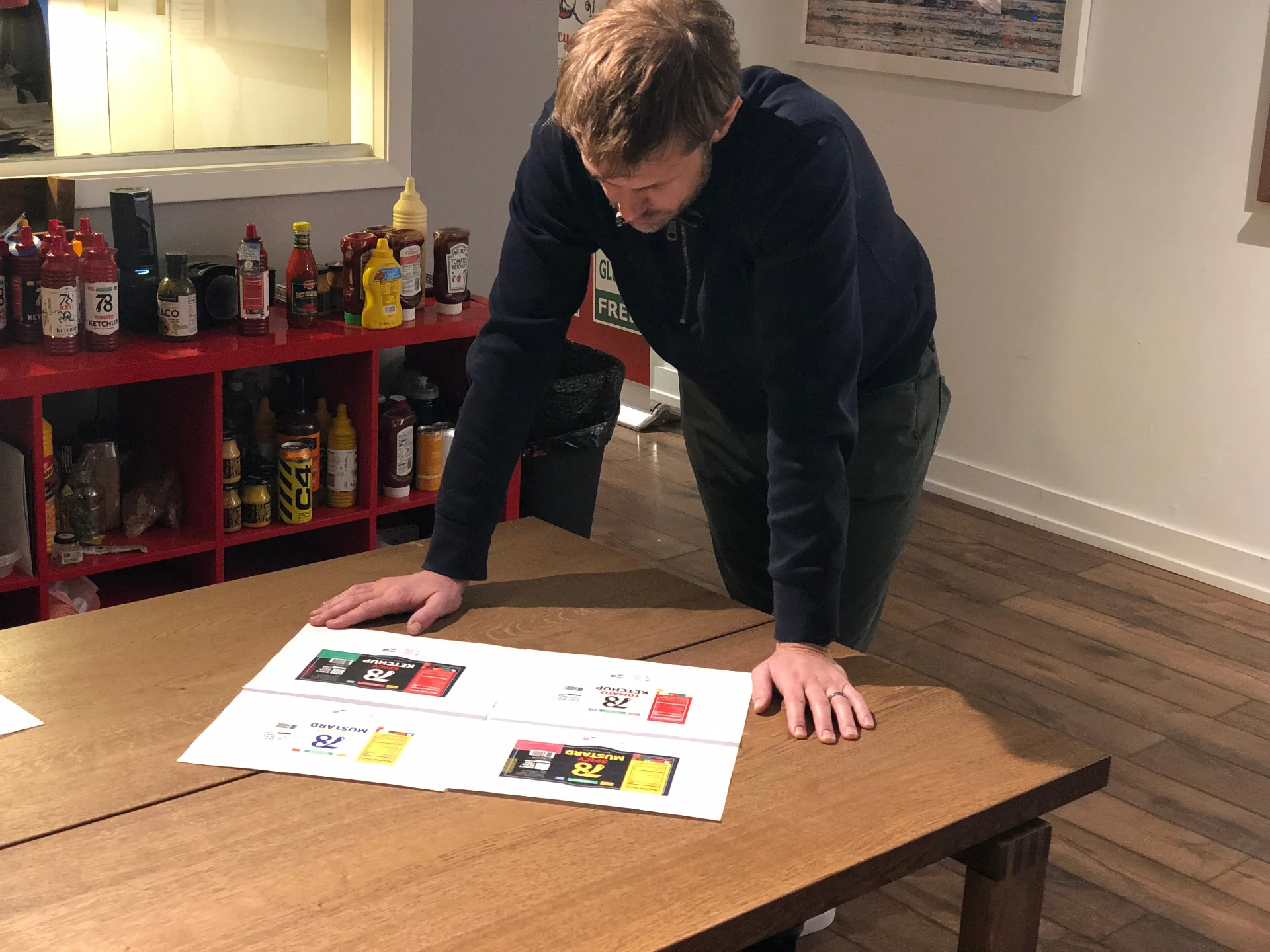 78 Brand founder reviewing printed ketchup and mustard label proofs against a shelf of competitor bottles